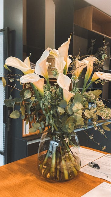 A tall, elegant floral arrangement featuring large, creamy white calla lilies with smooth, trumpet-shaped petals and prominent yellow centers. The lilies are complemented by sprigs of delicate white b