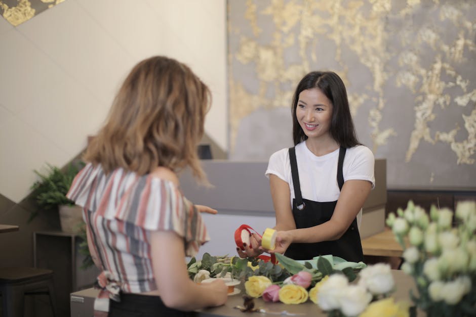 A professional florist with long dark hair, wearing a white t-shirt and black apron, smiling while handing a small floral gift wrapped with a yellow ribbon and red tape to a female customer with shoul