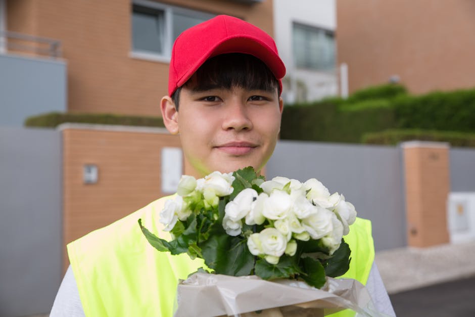 A young delivery person wearing a red cap and a neon yellow vest is holding a bouquet of white flowers, which appear to be miniature roses or similar blooms, wrapped in light-colored paper. The bouque