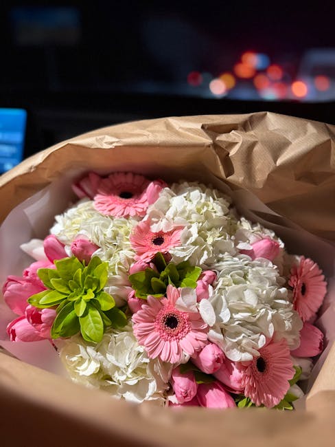 A bouquet of fresh flowers arranged in a brown paper wrapper, featuring white hydrangeas with delicate, ruffled petals, complemented by vibrant pink gerbera daisies showcasing dark central discs and l
