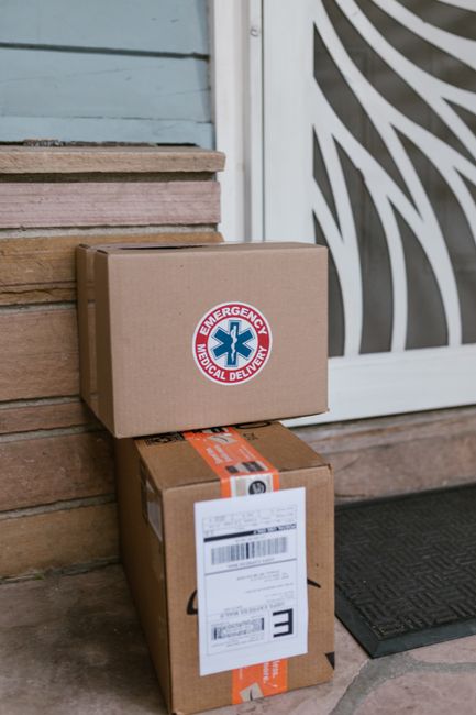 Two brown cardboard delivery boxes are stacked outside a building, with the top box displaying an emergency medical delivery sticker featuring a blue star of life symbol and red lettering. The boxes a