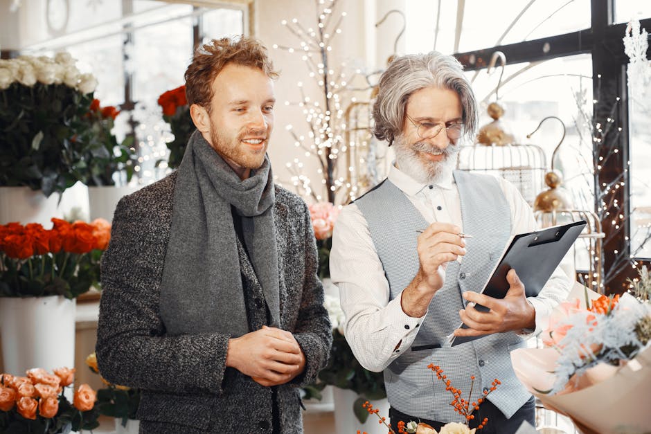 A young man with light brown hair and a grey scarf, dressed in a dark grey textured coat, stands beside an older man with grey hair, a beard, and glasses, wearing a light grey waistcoat over a white s